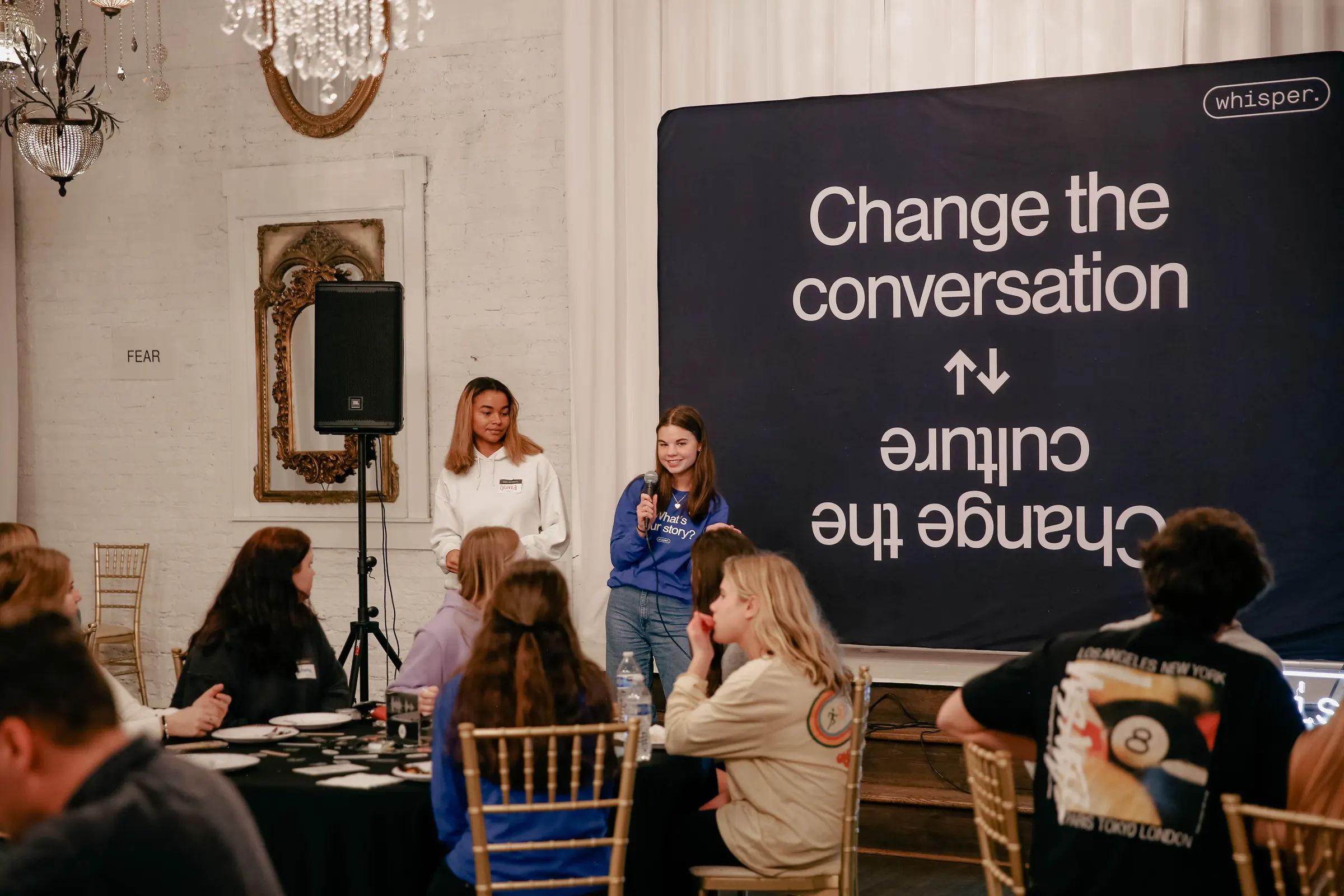 Students at round tables under chandeliers at a Whisper gathering, with a “Connection changes everything” screen